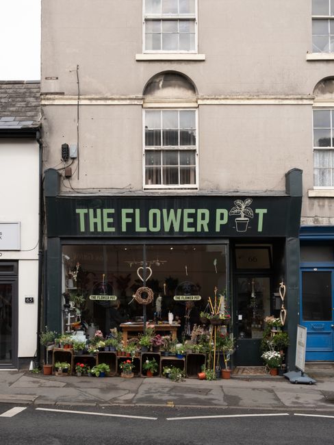 The storefront of The Flower Pot, a flower shop on Brent Street Hendon, featuring a large black sign with green lettering and a small flower pot logo. The display window showcases an array of fresh fl